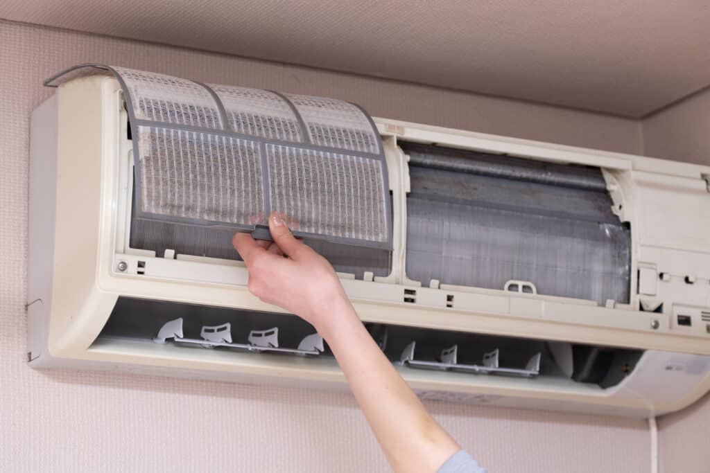 Main retirant un filtre à poussière encrassé d'un climatiseur mural blanc, révélant les ailettes sales à l'intérieur. Entretien.