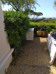 Allée de jardin en dalles de galets avec buissons verts, meuble extérieur et plantes en pots. Vue sur haie, arbres et mer bleue.
