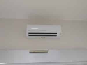 Climatiseur mural blanc Mitsubishi Electric allumé, installé sur un mur clair. Une petite barre beige est fixée en dessous.