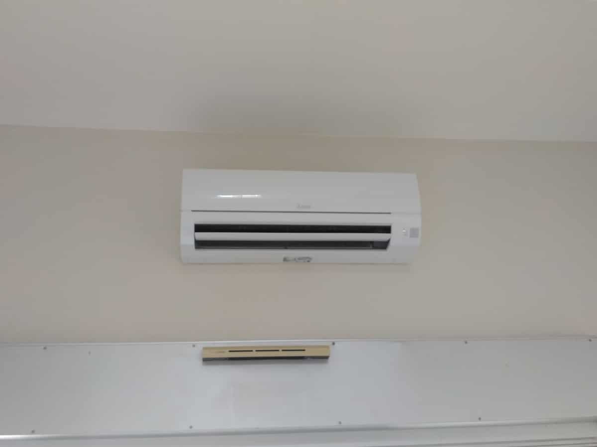 Climatiseur mural blanc Mitsubishi Electric allumé, installé sur un mur clair. Une petite barre beige est fixée en dessous.