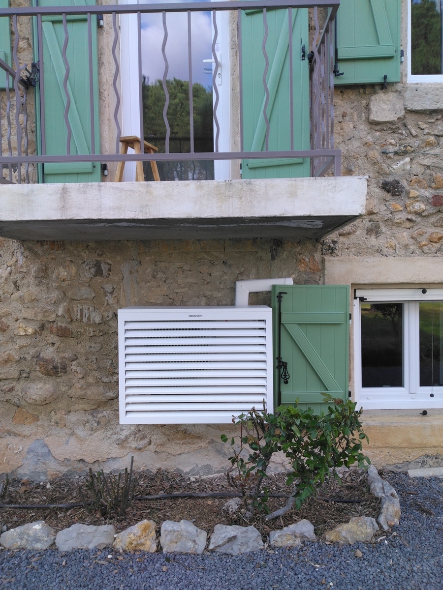 Maison en pierre avec balcon et volets verts. Un boîtier DECOClim est sur le mur sous la porte vitrée. Jardin avec gravier au premier plan.